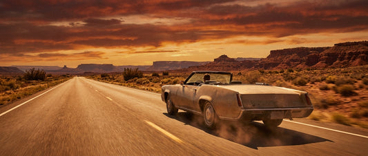 A vintage car driving along a straight desert highway toward a glowing amber horizon, flanked by red rock mesas under a dramatic open sky.
